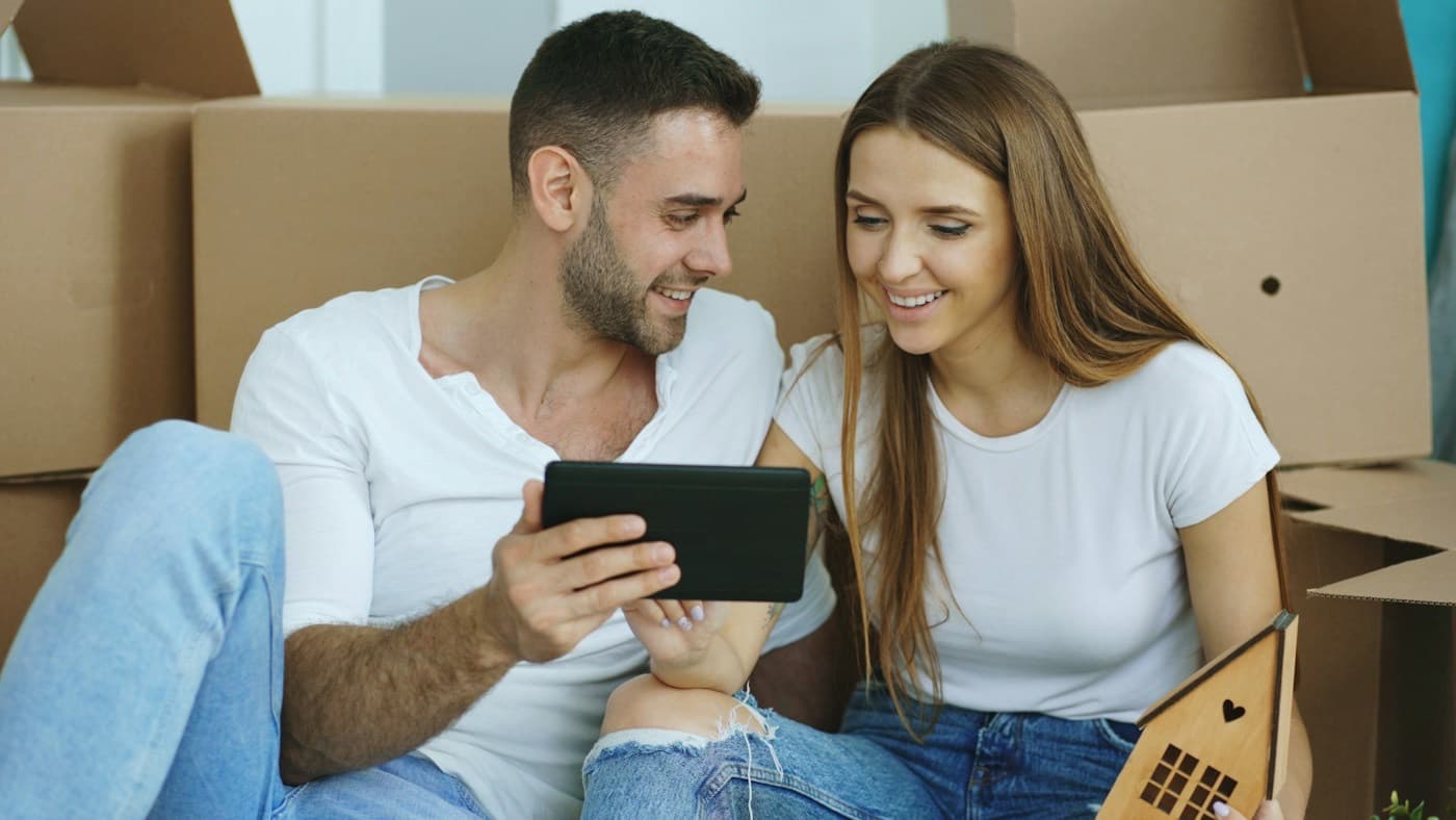 Casal sorrindo com um tablet nas mãos, sentado entre caixas de mudança