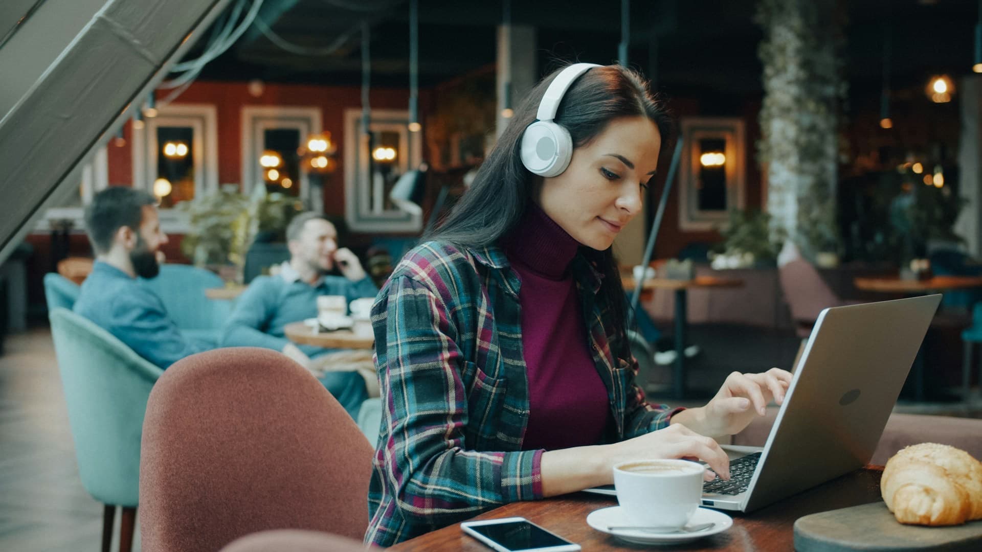 Mulher de fones de ouvido trabalhando no notebook em uma cafeteria, com o celular sobre a mesa